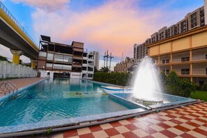 A swimming pool with a fountain, surrounded by buildings and a bridge. - Aristro Club (Alipur)