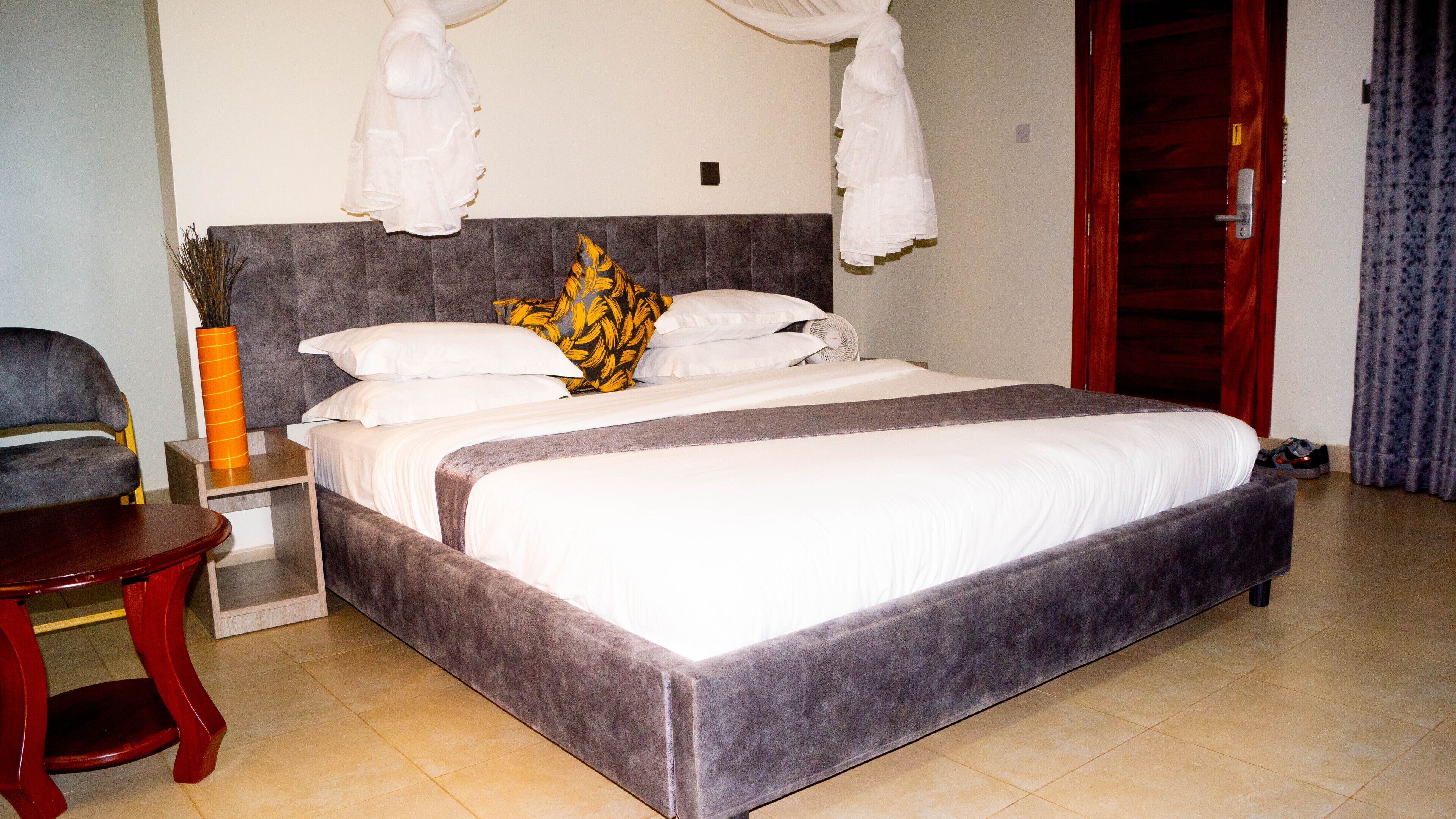 Una cama bien hecha con un cabecero gris, ropa de cama blanca y una almohada decorativa amarilla y negra. Junto a la cama hay una mesita de noche de madera con un florero de plantas secas. Una silla de madera es visible en primer plano.