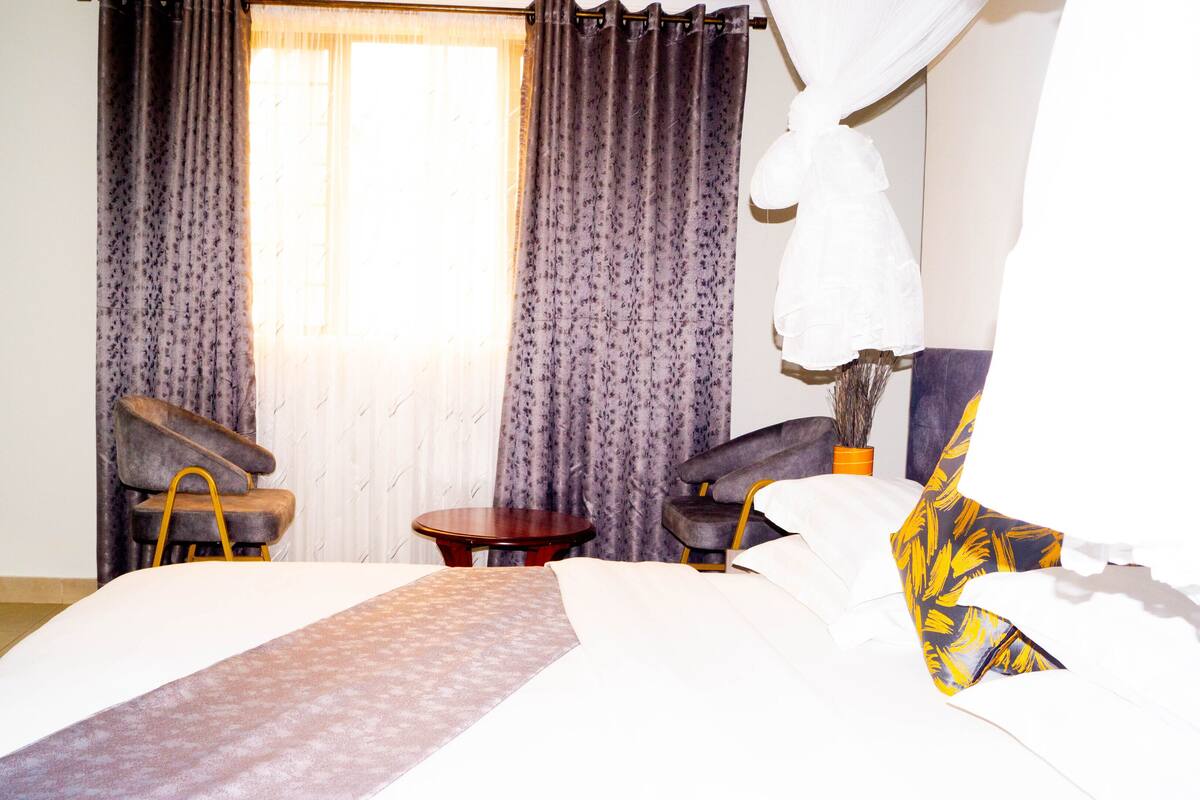 Quarto de hotel com uma cama, duas poltronas, uma mesinha redonda e uma cortina.