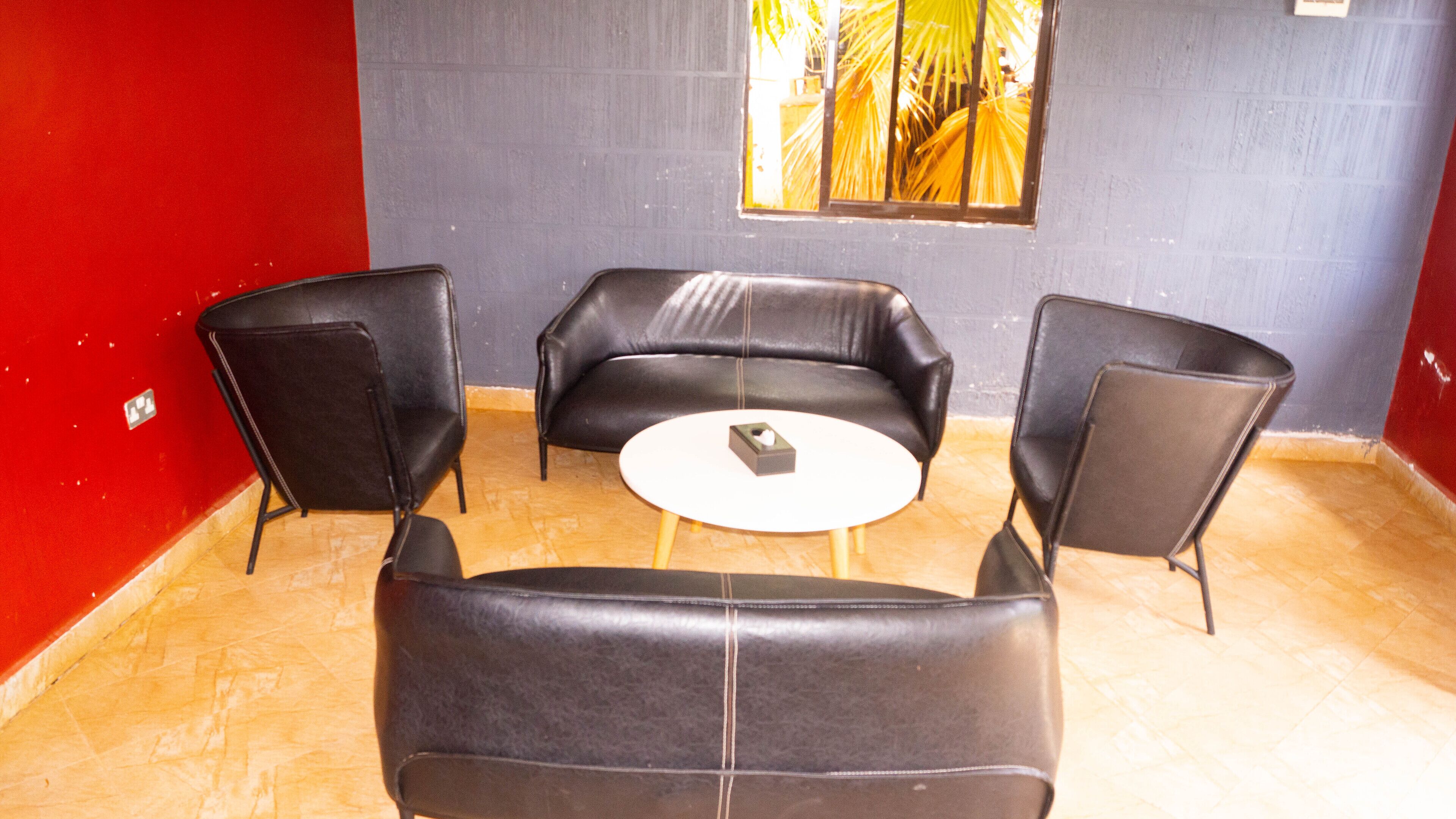 Habitación con una pared roja, una pared gris con ventana y suelo de madera. Hay un sofá de cuero negro, un sillón de cuero negro y una mesa redonda blanca con un objeto pequeño encima.
