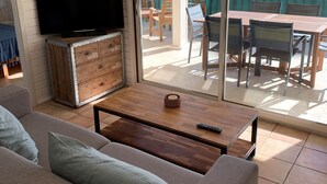 A living room with a sofa, a coffee table, and a television on a wooden cabinet. There is a sliding glass door leading to an outdoor patio with a table and chairs.