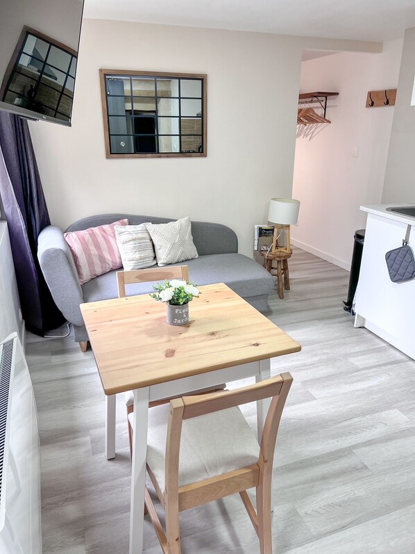 Un salon moderne avec un canapé gris, une table à manger en bois avec une chaise, une petite table d’appoint avec une lampe et une fenêtre donnant sur l’extérieur.