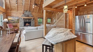 Interior de una cabaña de troncos con chimenea de piedra, un comedor, una cocina con electrodomésticos de acero inoxidable y una escalera que conduce a un nivel superior.