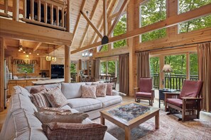 A spacious living area with a large sofa, a coffee table, and two armchairs. There is a kitchen area with wooden cabinets and a dining table with chairs. Large windows provide a view of the outdoors. - Luxury 4 BR with Hot Tub, Seclusion, WiFi, Huge Decks, Free Dollywood Tickets (Sevierville)