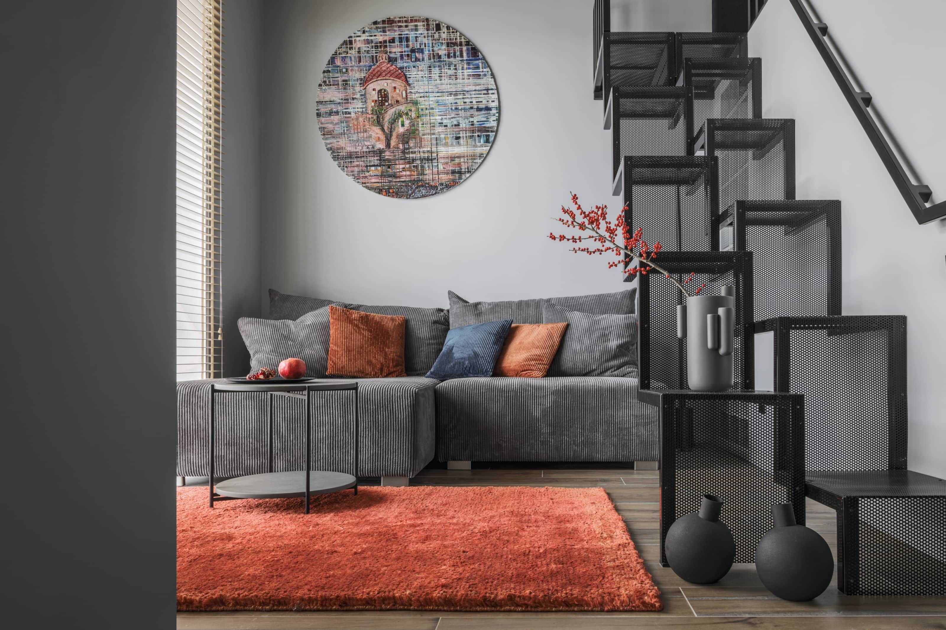 A modern living room with a grey sofa, a round wall art piece, a staircase, and a red rug.