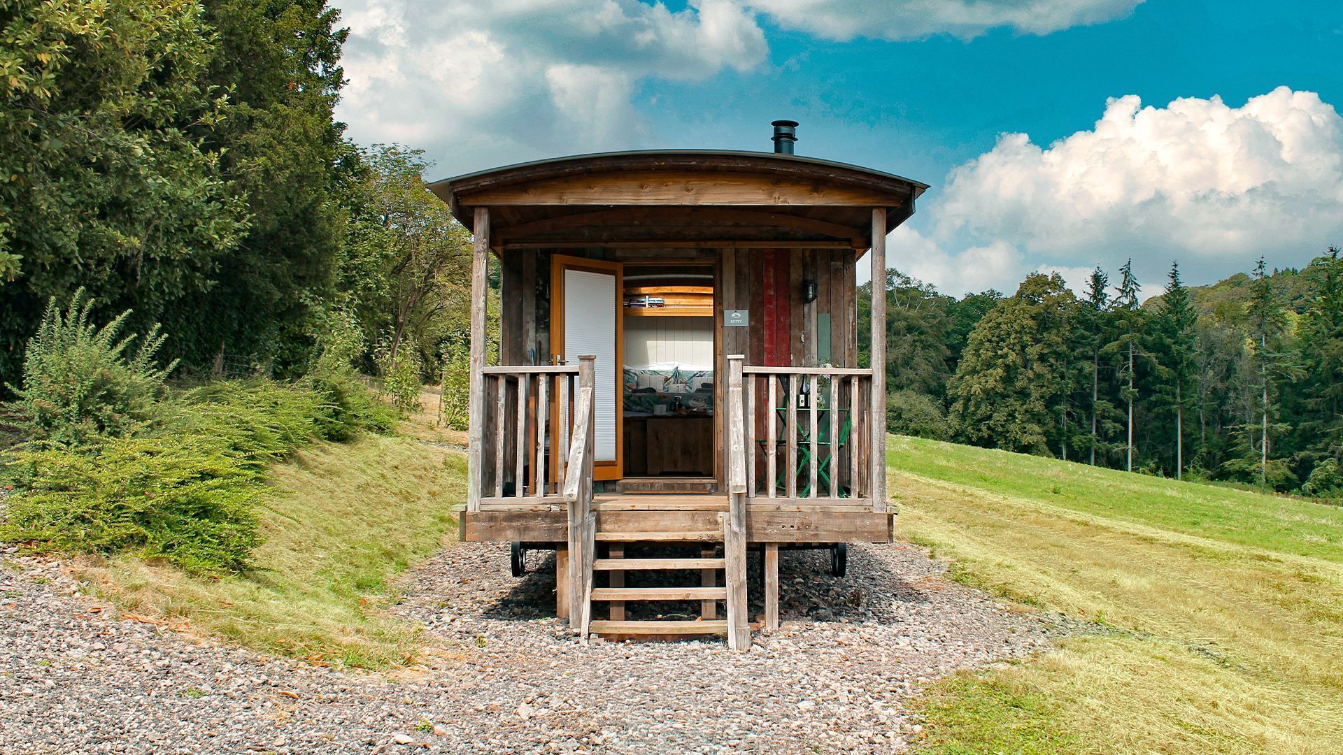 A wooden cabin with a balcony, surrounded by trees and a gravel path.