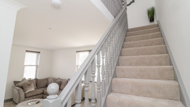 Un escalier avec des marches recouvertes d’une moquette beige et des balustrades blanches, menant à un palier doté d’un plafond blanc et d’un détecteur de fumée.