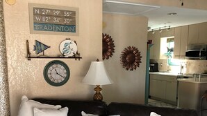 Een bruine leren bank met witte kussens, een wandklok, decoratieve wandkunst en een keuken op de achtergrond.