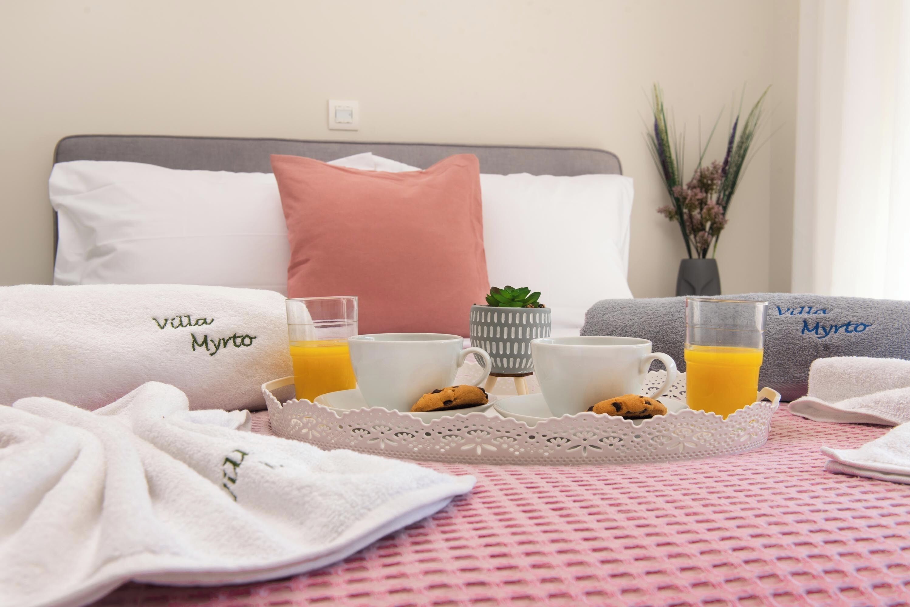 Uma mesa de café da manhã bem organizada, com suco de laranja, biscoitos e xícaras de café, toalhas com a marca 'Villa Myrto' e uma planta decorativa.