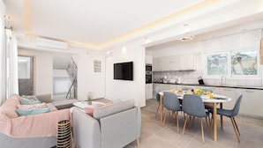 Un salon moderne comprenant un canapé, une table à manger avec des chaises, un téléviseur fixé au mur et un coin cuisine avec des placards et une fenêtre.