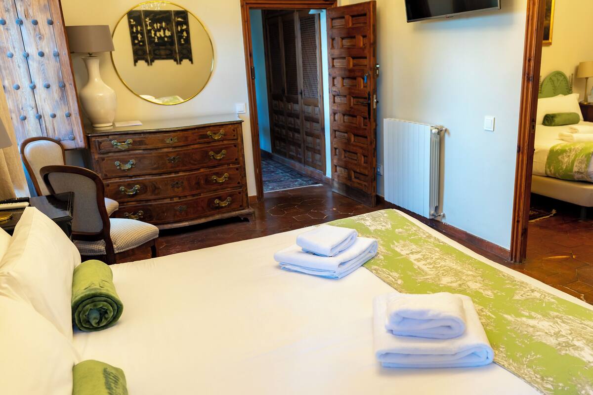 Une chambre d’hôtel avec un lit, une commode en bois, une chaise, un bureau avec une lampe, un téléviseur et une salle de bain visible à travers une porte ouverte.