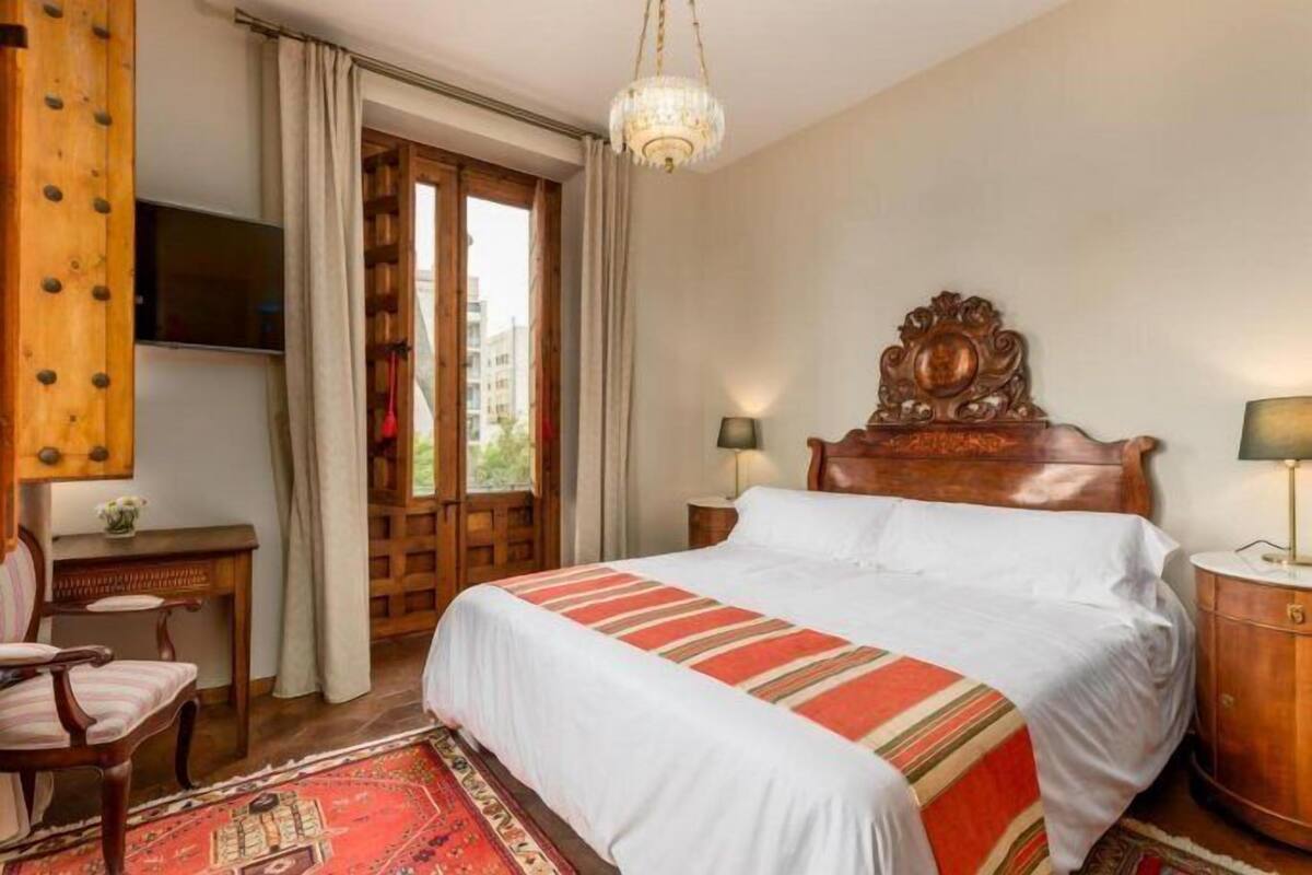 Une chambre d’hôtel avec un grand lit, une tête de lit en bois, deux tables de chevet avec des lampes, un téléviseur fixé au mur, un bureau en bois et un tapis rouge à motifs.