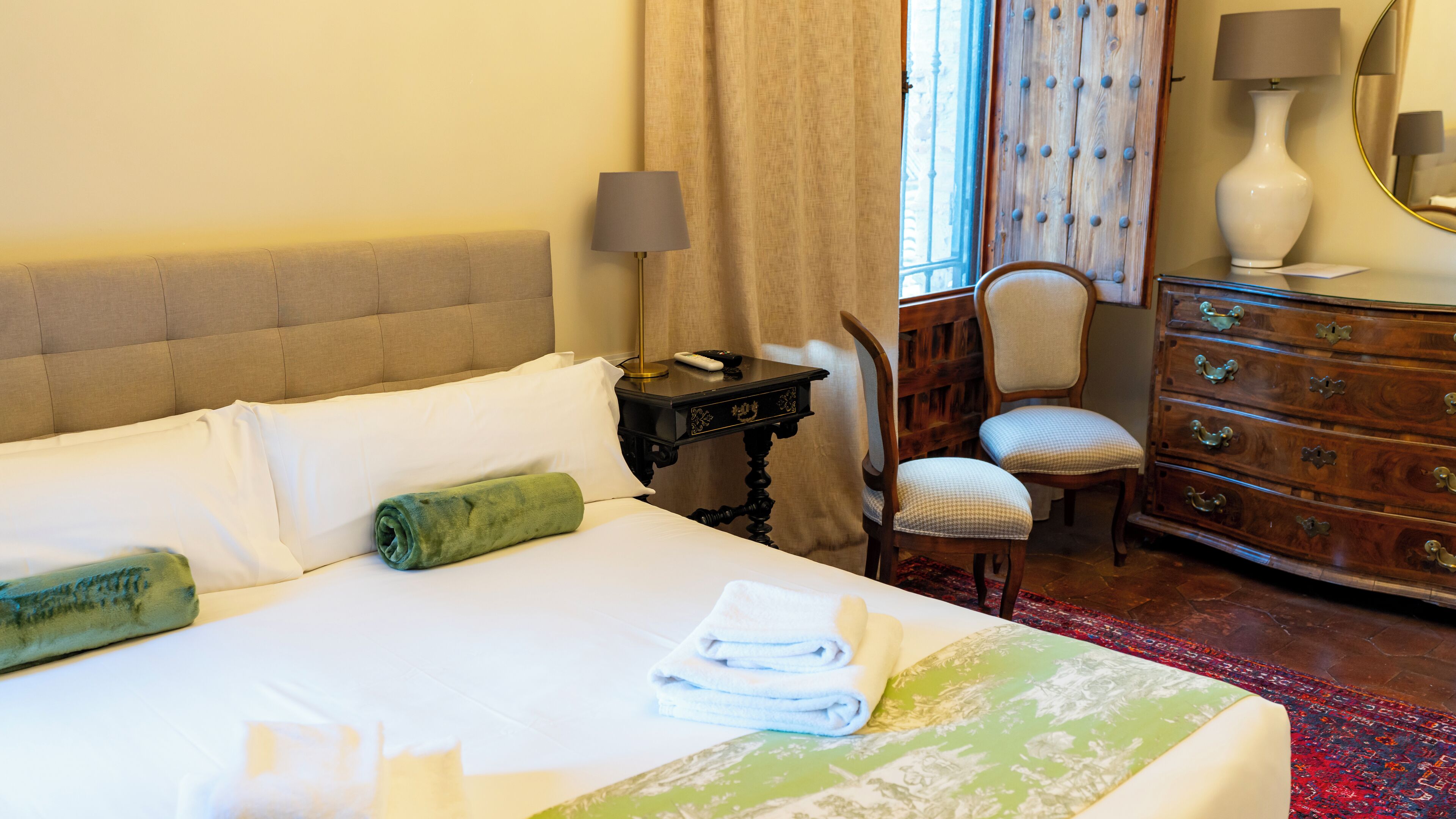 A neatly made bed with white linens, a green towel, and a folded white towel. There is a wooden nightstand with a lamp and a remote control. A wooden dresser with drawers and a mirror is visible. A chair with a checkered cushion is placed next to the dresser.