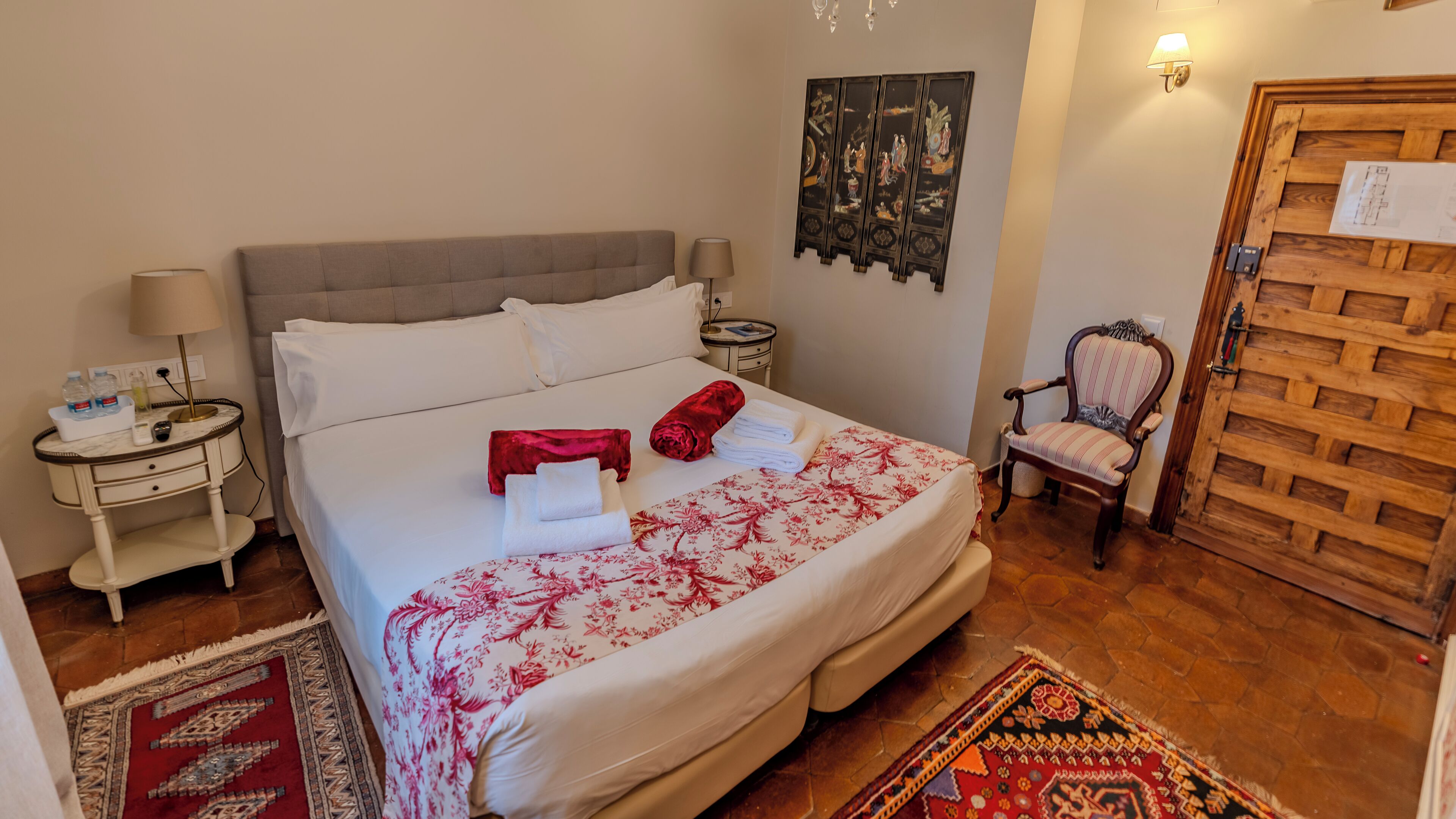 A neatly made bed with a floral bedspread, two bedside tables with lamps, a chair, and a wooden door with a latch.