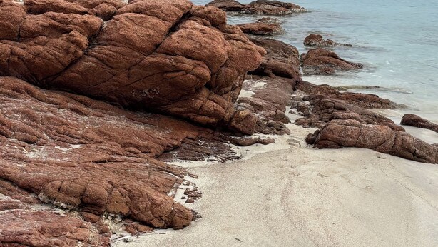 A rocky shoreline with clear water, a sandy beach, and distant buildings.
