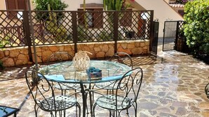 A glass-top table with metal chairs, a stone floor, and a metal fence in a sunny outdoor area.
