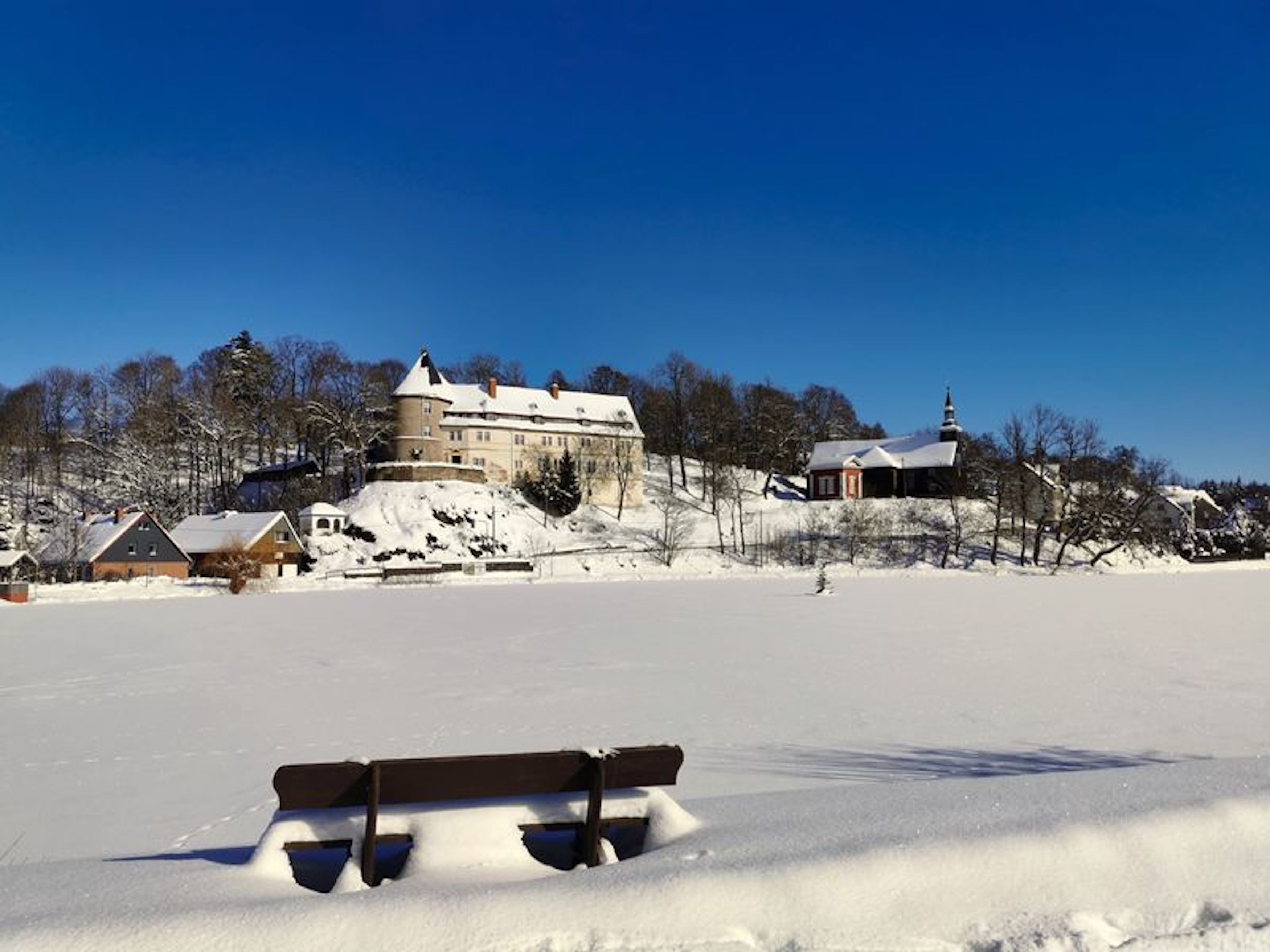 Een besneeuwd landschap met een groot gebouw op een heuvel, een kleinere structuur met een torenspits en een bank op de voorgrond.