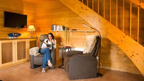 L’intérieur chaleureux d’un chalet avec un escalier en bois, un téléviseur fixé au mur et un coin salon comprenant un fauteuil et une table.