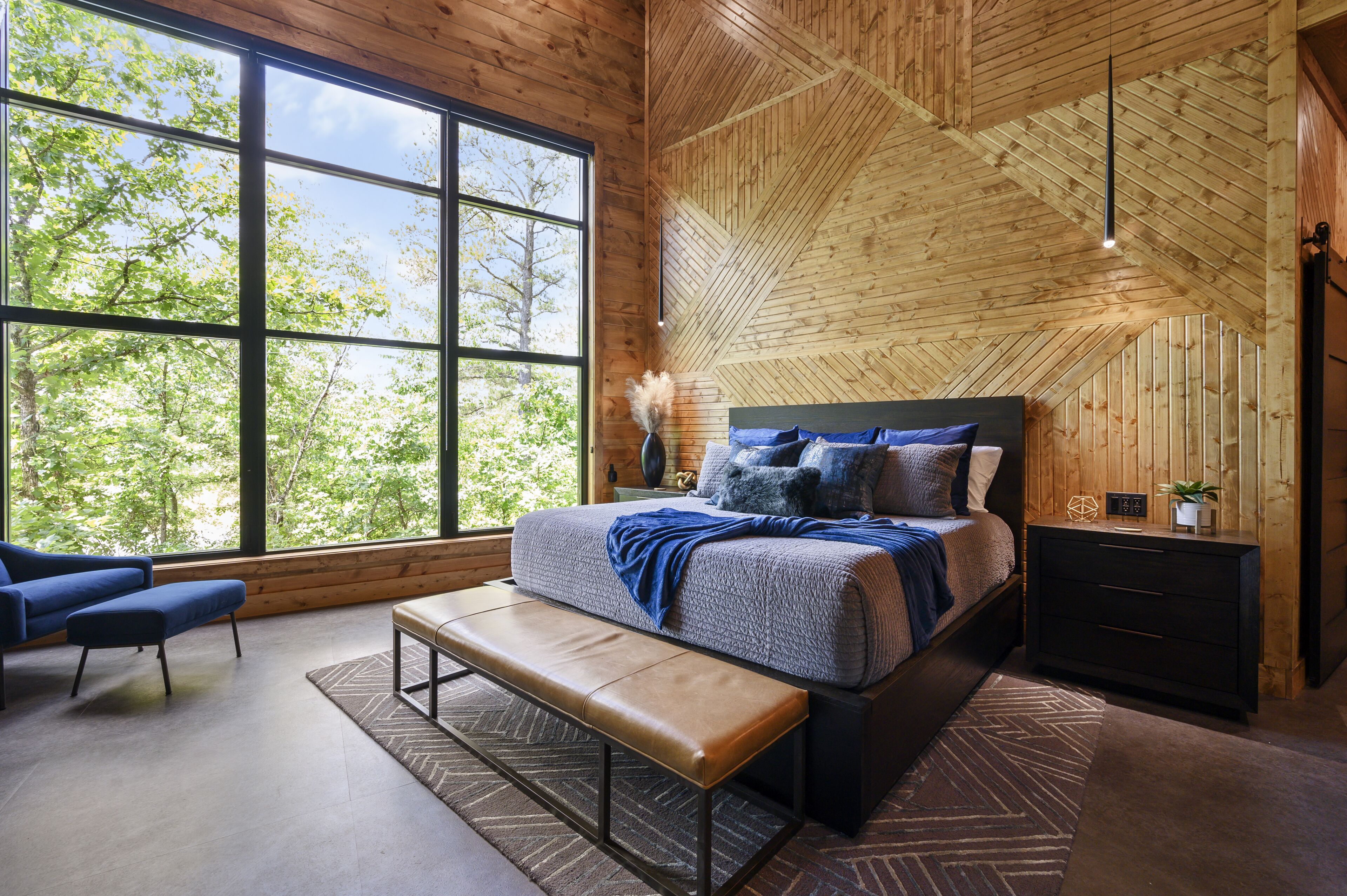A bedroom with a large window, a bed, a bench, a nightstand, and a dresser.