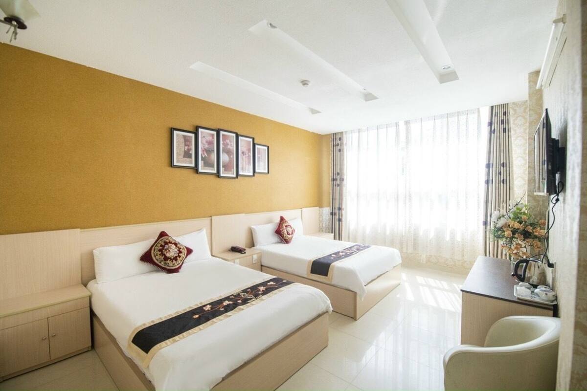 A hotel room with two beds, a desk, and a chair. There are framed pictures on the wall and a window with curtains.