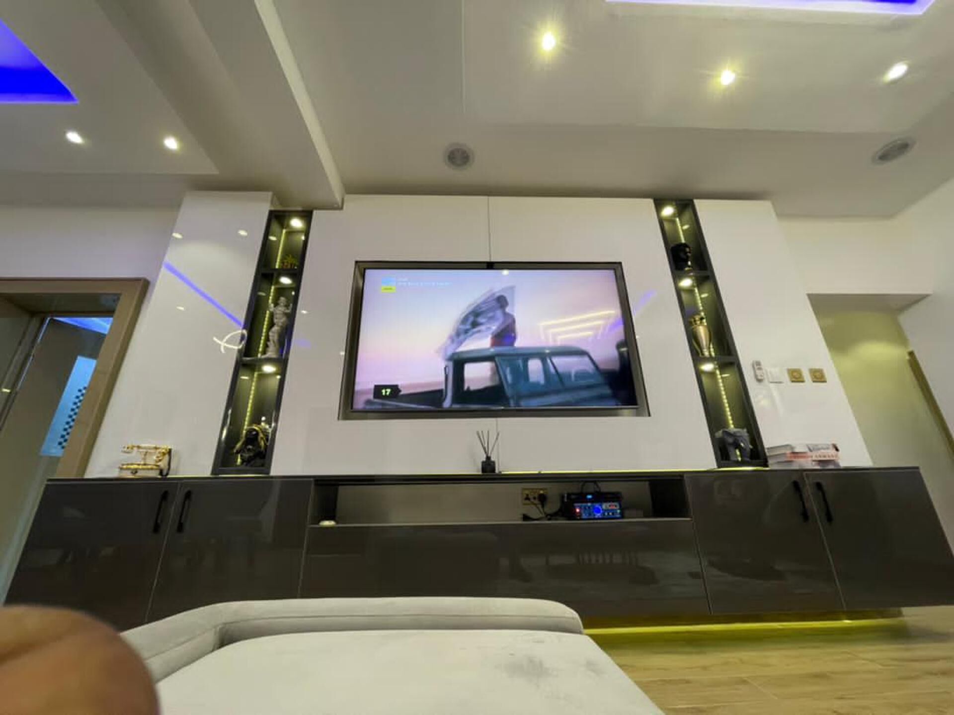 Un salon moderne doté d’un grand téléviseur à écran plat fixé au mur, d’un meuble de rangement de couleur sombre placé en dessous, et d’un plafond encastré avec un éclairage intégré.
