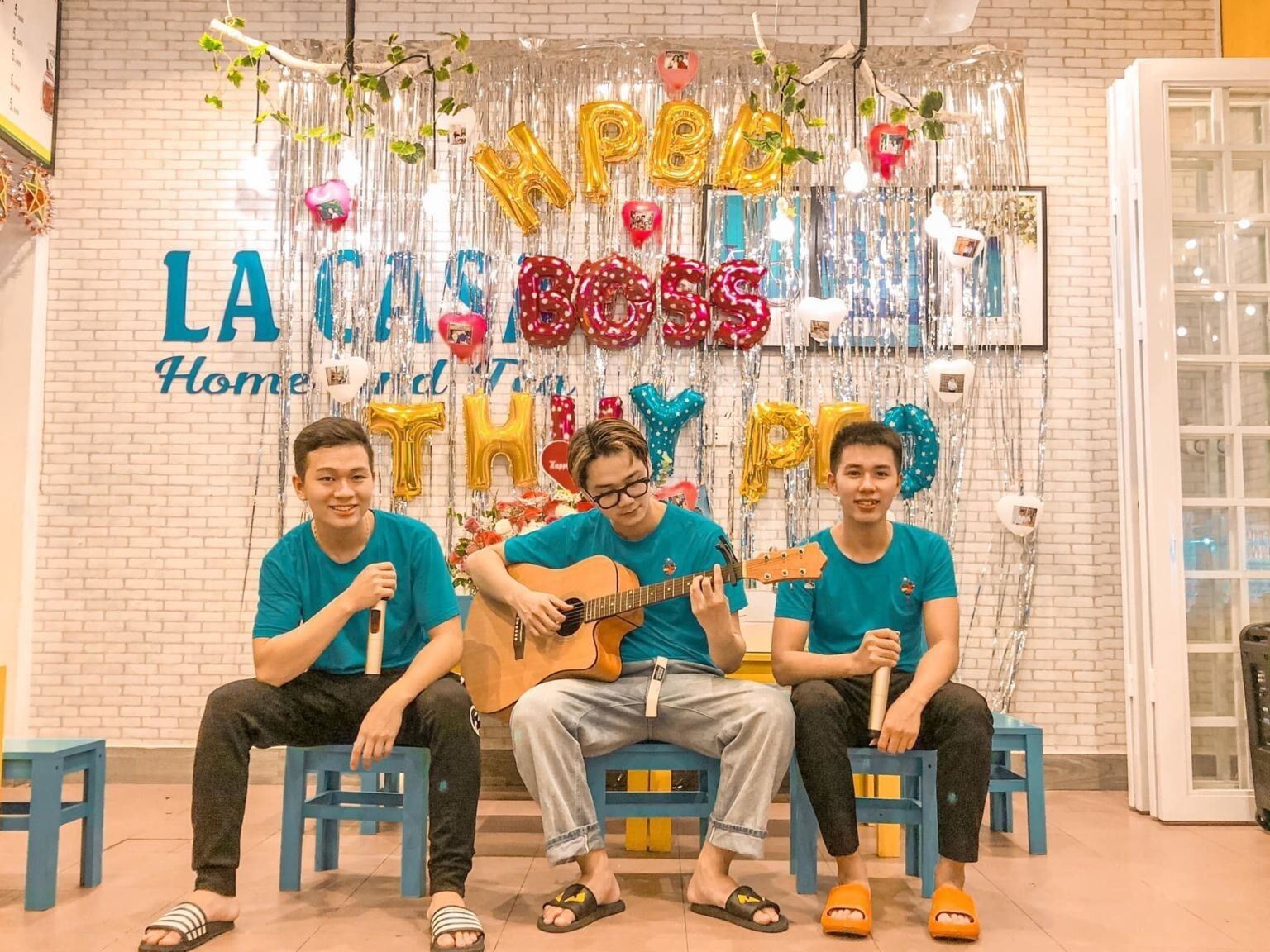 Three people sitting on blue chairs in front of a wall decorated with balloons and the text "Happy Birthday".