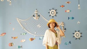 A room with a blue wall decorated with nautical elements, a yellow bench, and a person in a green dress and hat. - La Casa Homestay Ha Long (Bai Chay)