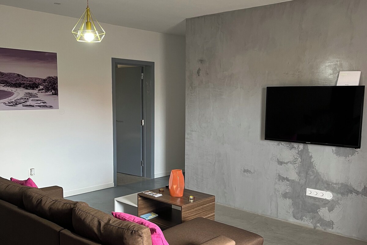 A modern living room with a sectional sofa, a flat-screen TV mounted on a textured wall, a pendant light, and a door leading to another room.