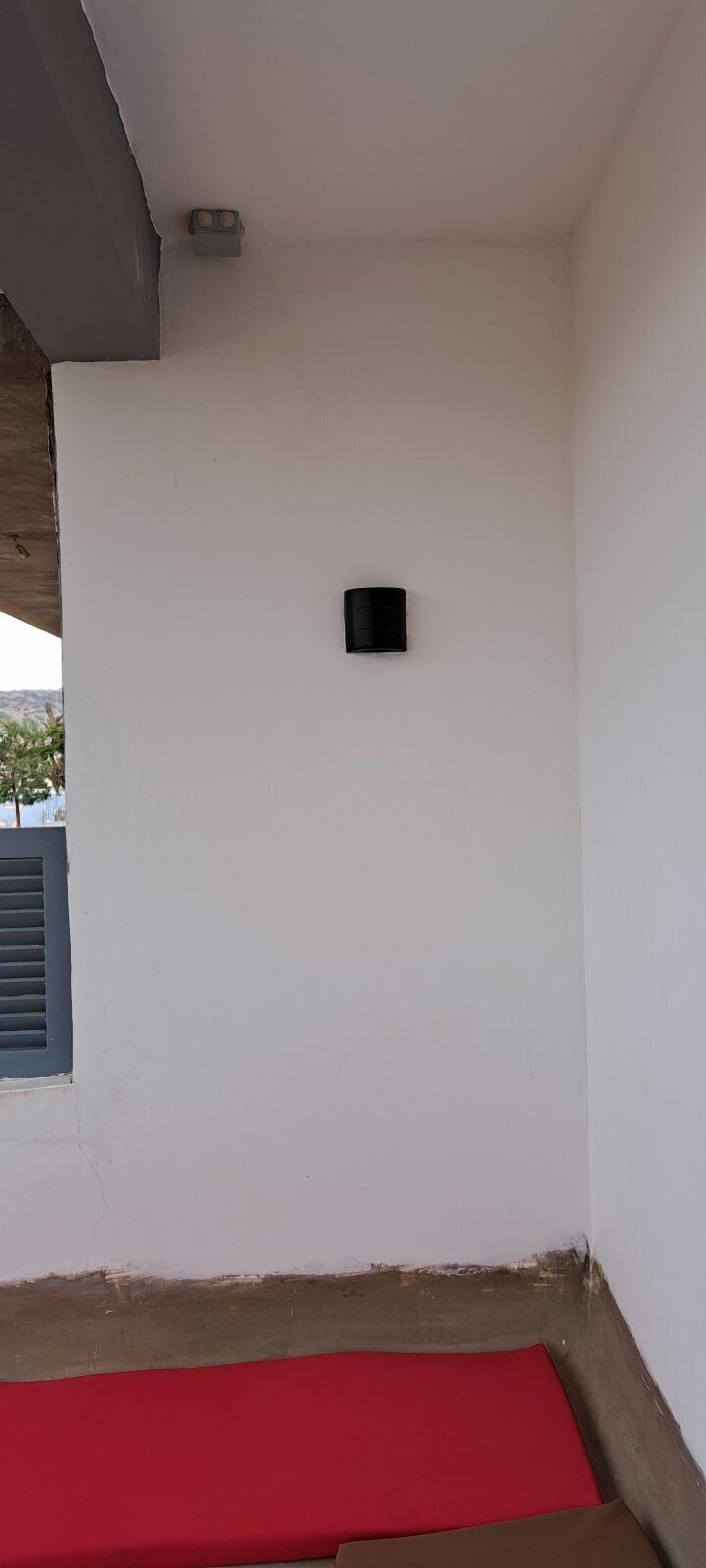 A room with a white wall, a small black object on the wall, and a red mat on the floor.