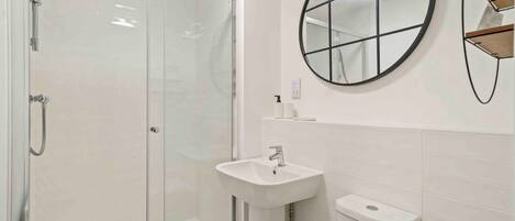 A bathroom with a glass-enclosed shower, a white sink, a toilet, and a round mirror on the wall.