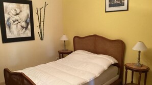 Un lit simple avec un cadre en bois et une literie blanche, flanqué de deux tables de chevet avec des lampes, dans une pièce aux murs jaunes et ornée d’œuvres d’art au-dessus du lit.
