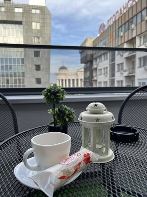 Et bord med en hvid kop, en lille potteplante, en hvid lygte og en snackpose på en balkon med udsigt over et bybillede med moderne bygninger.
