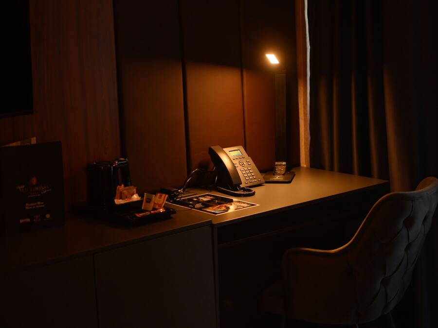Quarto de hotel com pouca luz, incluindo uma mesa, um telefone, uma cafeteira e uma cadeira.