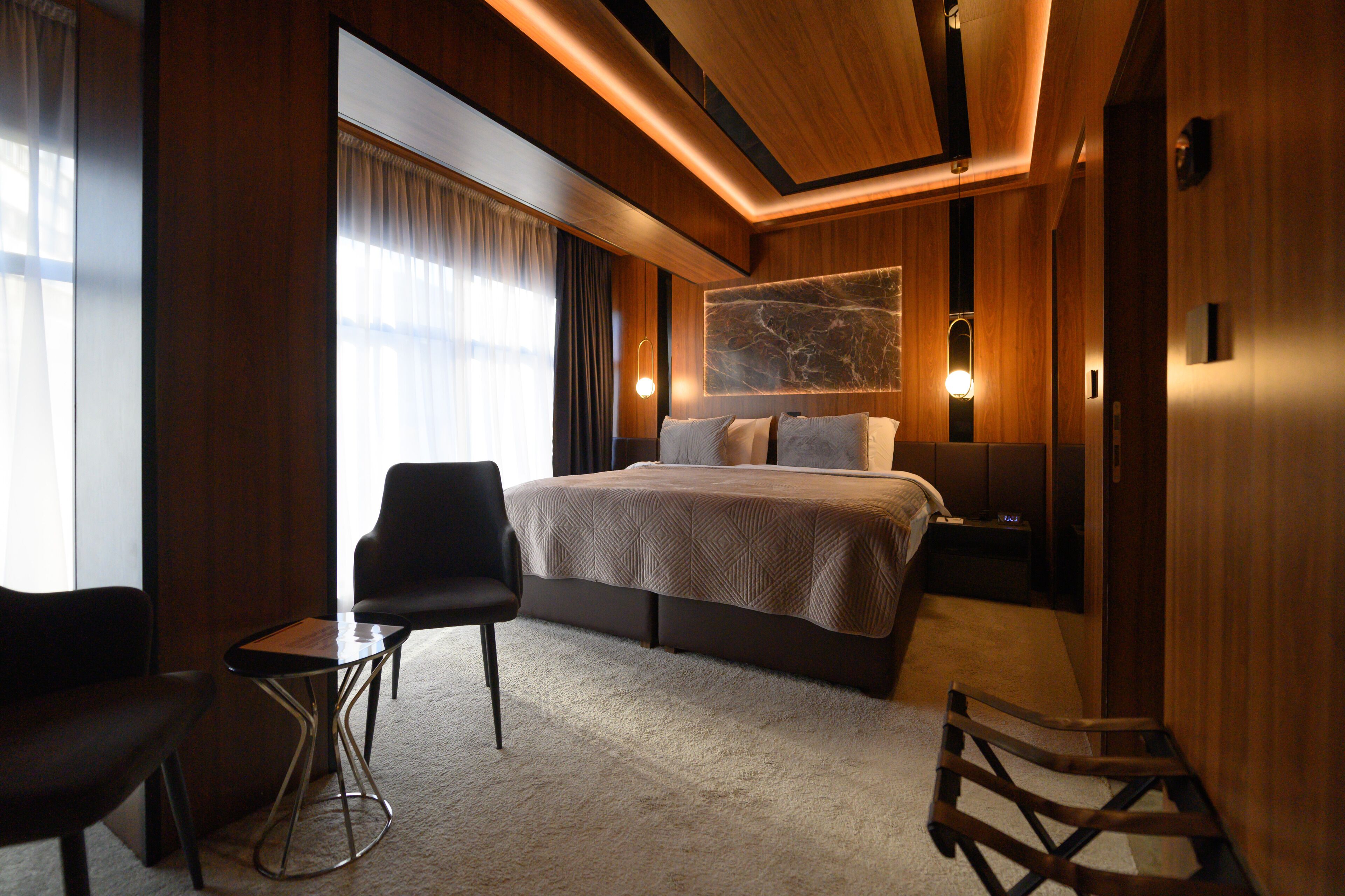 A modern hotel room with a large bed, a small table, two chairs, and a large window with curtains.