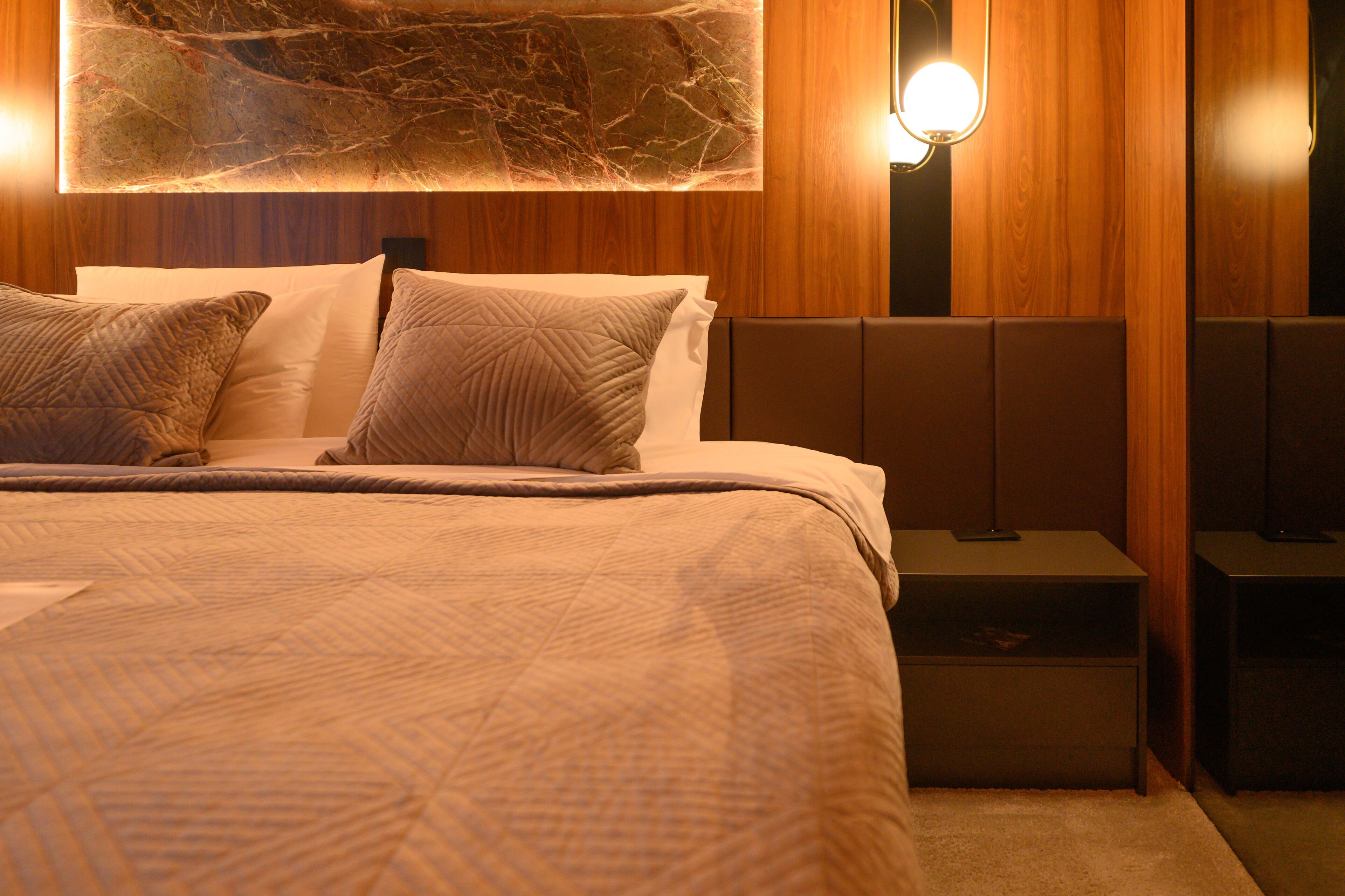 Un lit bien fait, avec une couvre-lit à motifs et des oreillers, une décoration murale en forme de marbre au-dessus, et une table de chevet avec une lampe.