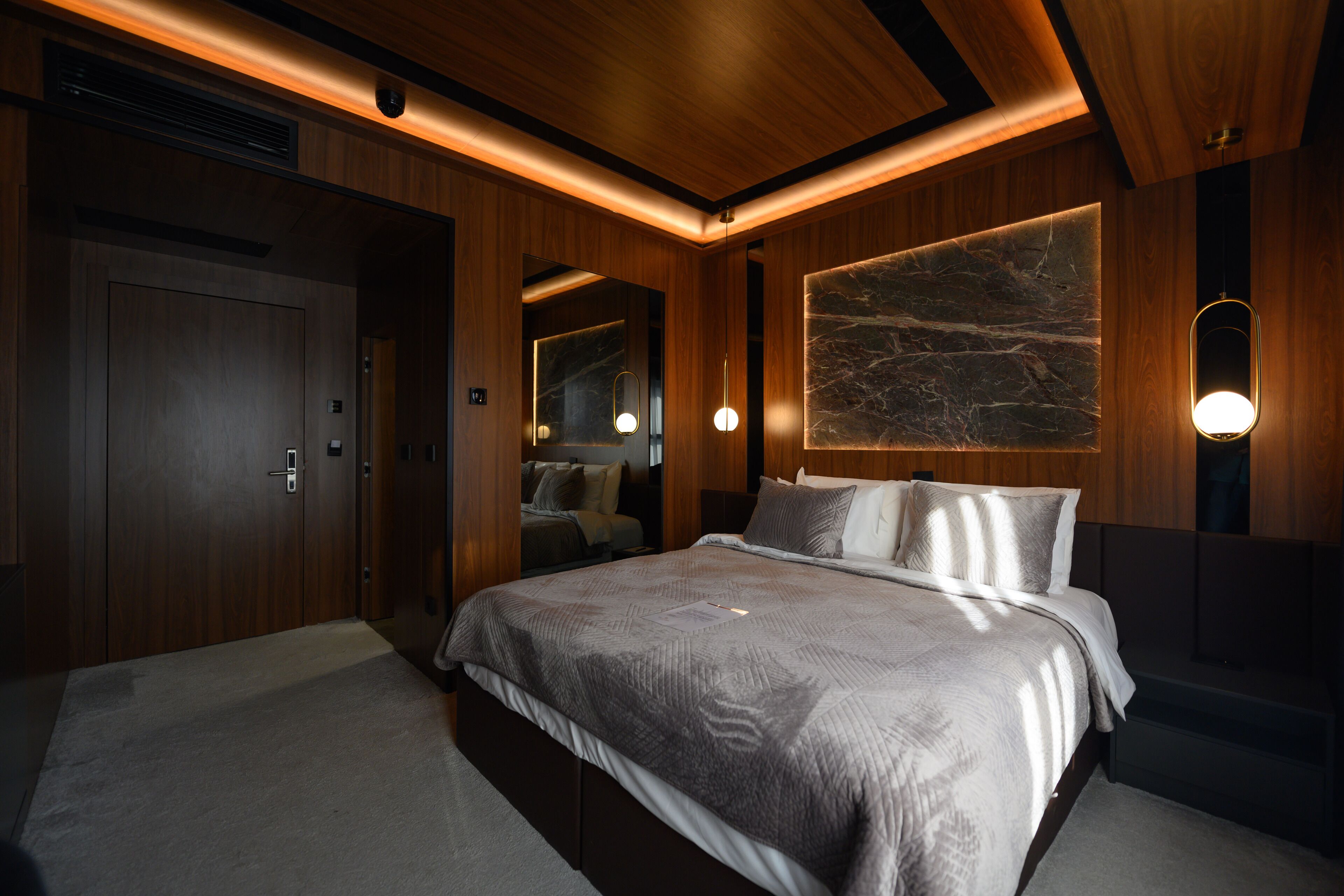 A modern bedroom with a large bed, wooden paneling, a marble accent wall, and pendant lighting.