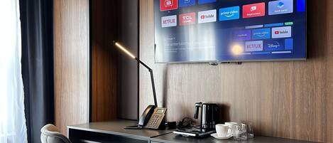 A modern hotel room with a large flat-screen TV displaying various streaming services, a desk with a telephone and coffee maker, and a comfortable chair.
