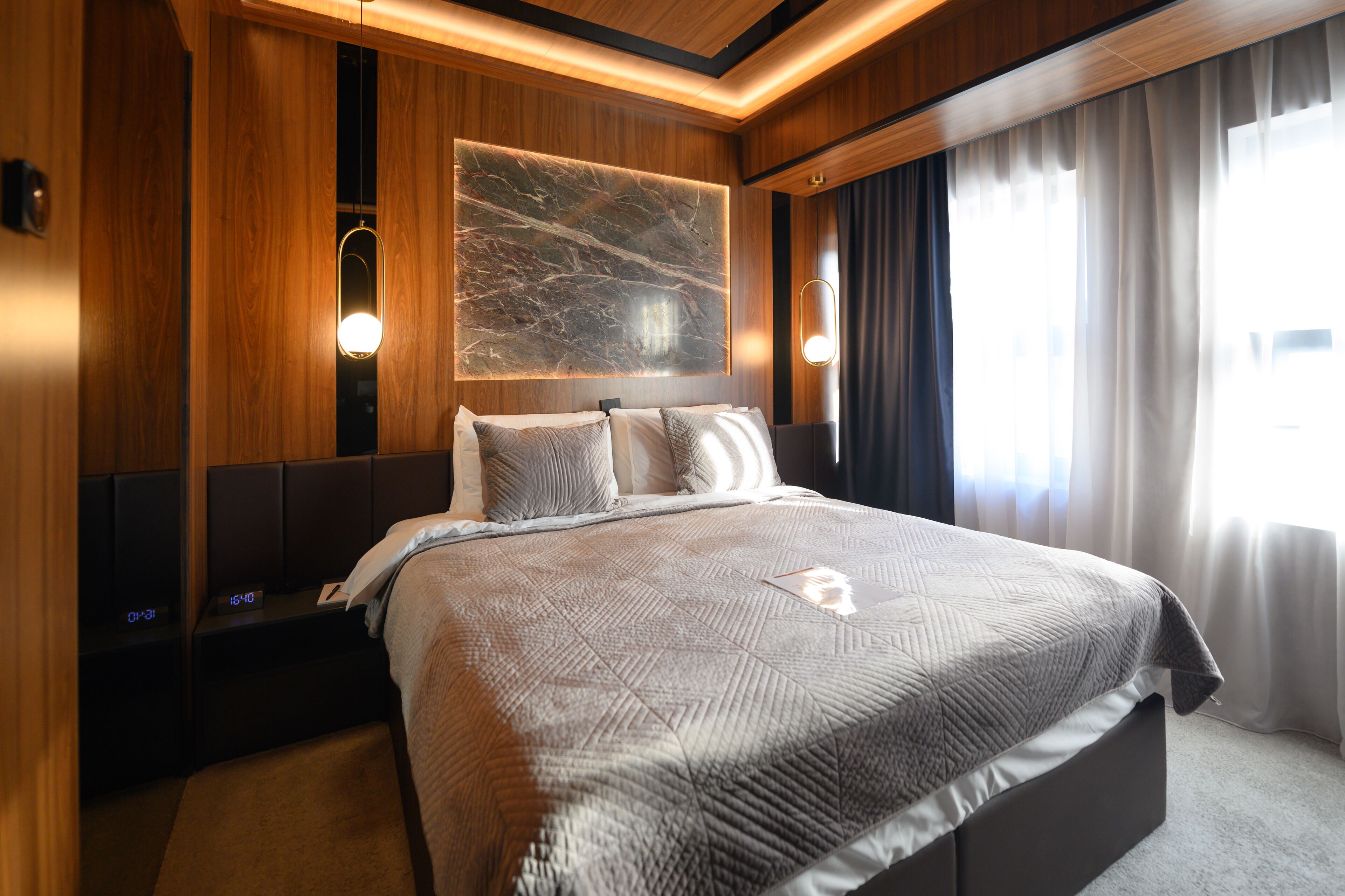 Habitación de hotel moderna con una cama grande, paneles de madera, una obra de arte de mármol en la pared y un amplio ventanal con cortinas.