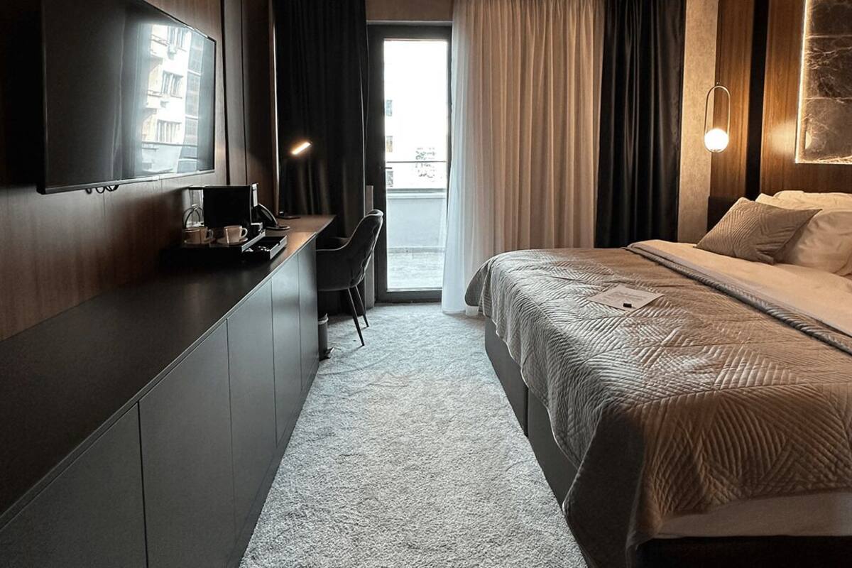 A modern hotel room with a large bed, a desk, a flat-screen TV, and a view of the city through the window.