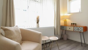 Een woonkamer met een beige bank, twee kleine witte tafels, een houten dressoir met getextureerde lades en een raam met gordijnen.