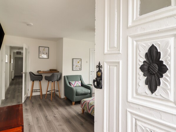 A modern living room with a grey sofa, two matching armchairs, a small table, and a colorful throw blanket. A white door with a black floral design is visible. - The Old Coach House (Conwy)