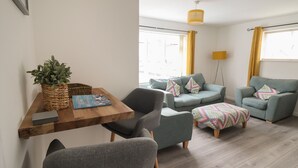 Een moderne woonkamer met een houten salontafel, grijze fauteuils, een bank en een gestreept voetenbankje. Op de tafel staat een potplant en er hangt een gele lamp.