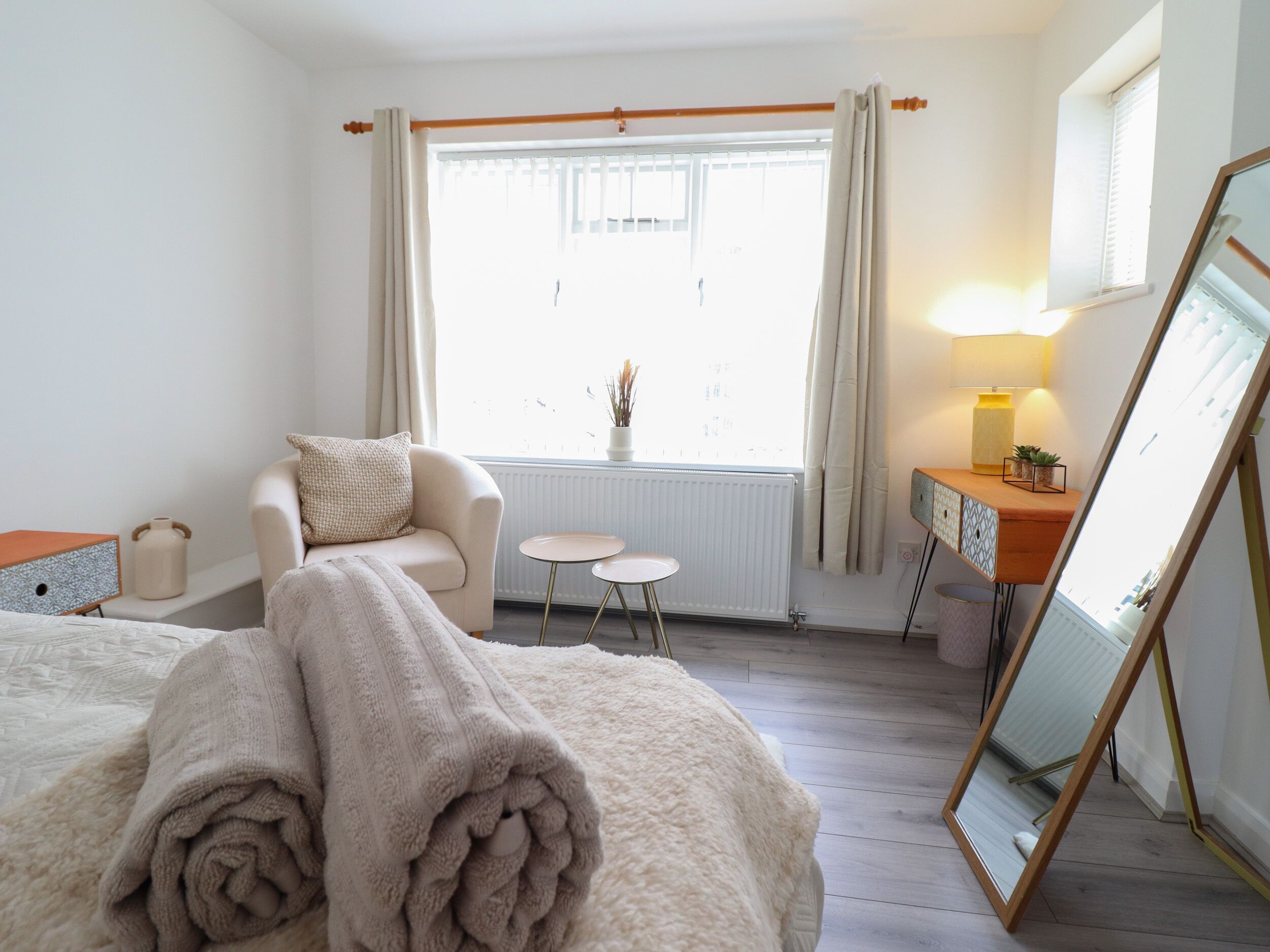 A bedroom with a bed, a chair, a small table, a dresser, a mirror, and a window with curtains.