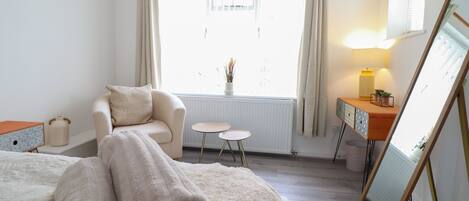 A bedroom with a bed, a chair, a small table, a dresser, a mirror, and a window with curtains.