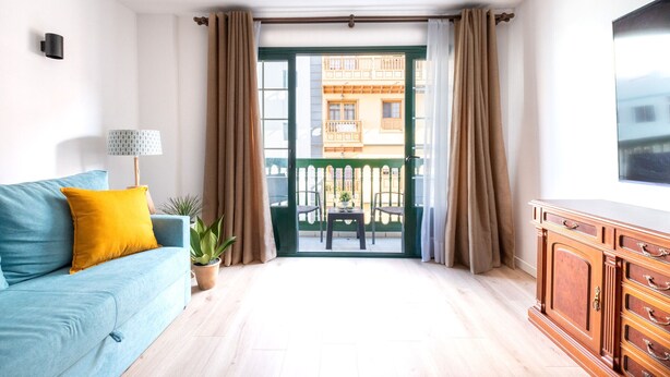 Et værelse med en blå sofa, et træskab, en potteplante og udsigt gennem en glasdør til en udendørs balkon.