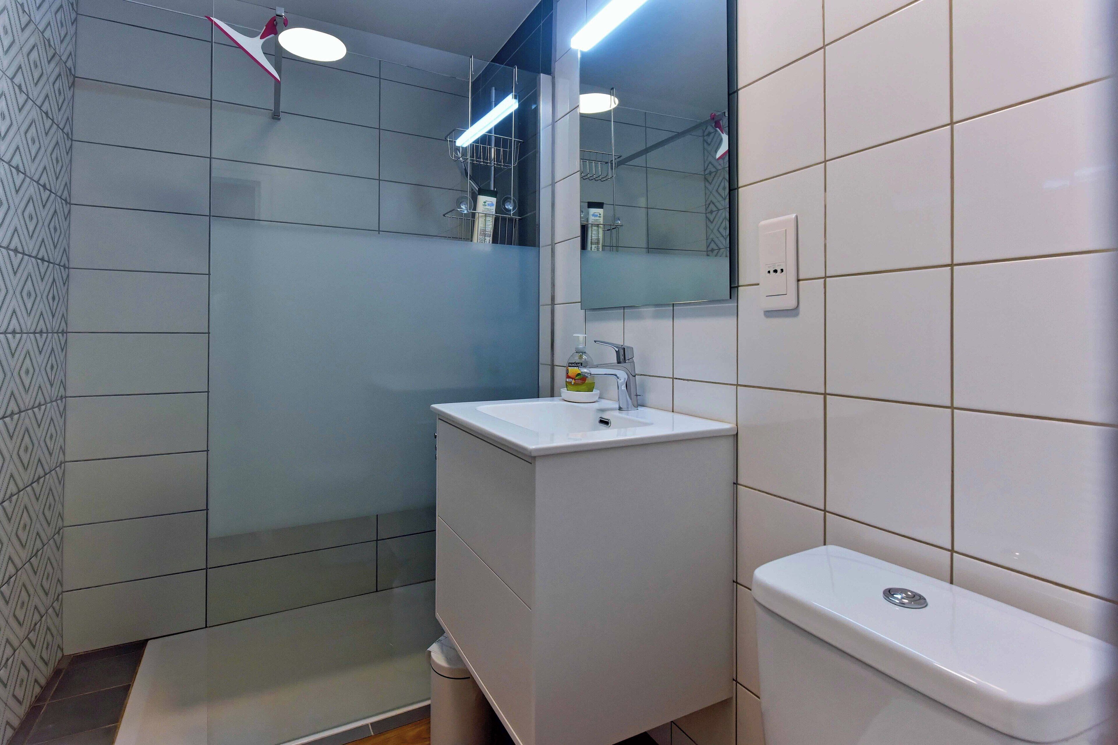 Moderni kylpyhuone, jossa on valkoinen pesuallas, peili ja wc-istuin. Kylpyhuoneessa on suihkutila, jossa on lasiovi, sekä pyyheteline.