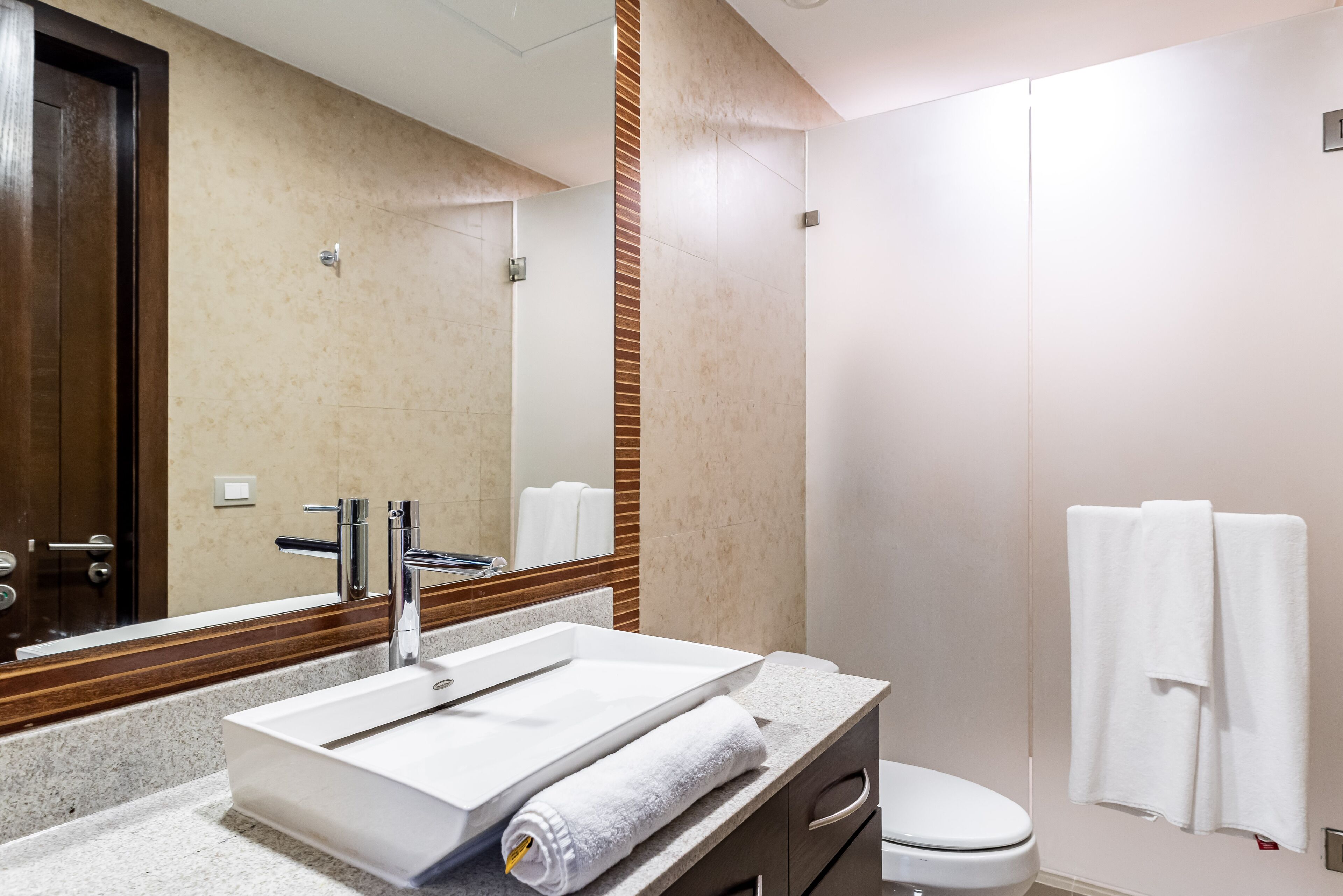 Une salle de bain moderne avec un grand miroir, un lavabo blanc, une serviette et des toilettes.