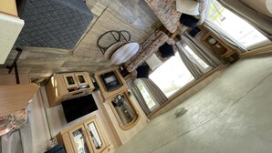 Un vaste salon comprenant un grand canapé, une table basse et un téléviseur. Il y a également un coin cuisine avec des meubles en bois et une table à manger. La pièce est dotée d’un haut plafond et de grandes fenêtres.