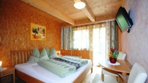 A room with a bed, a television mounted on the wall, a window with curtains, and a small table with a chair. - Truckshutte Holiday Home (Mayrhofen)