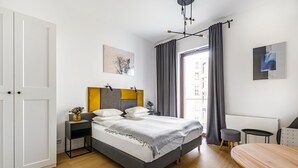 A modern bedroom with a bed, bedside tables, a small stool, and a dining table. - Homewell Apartments Żelazko (Poznan)